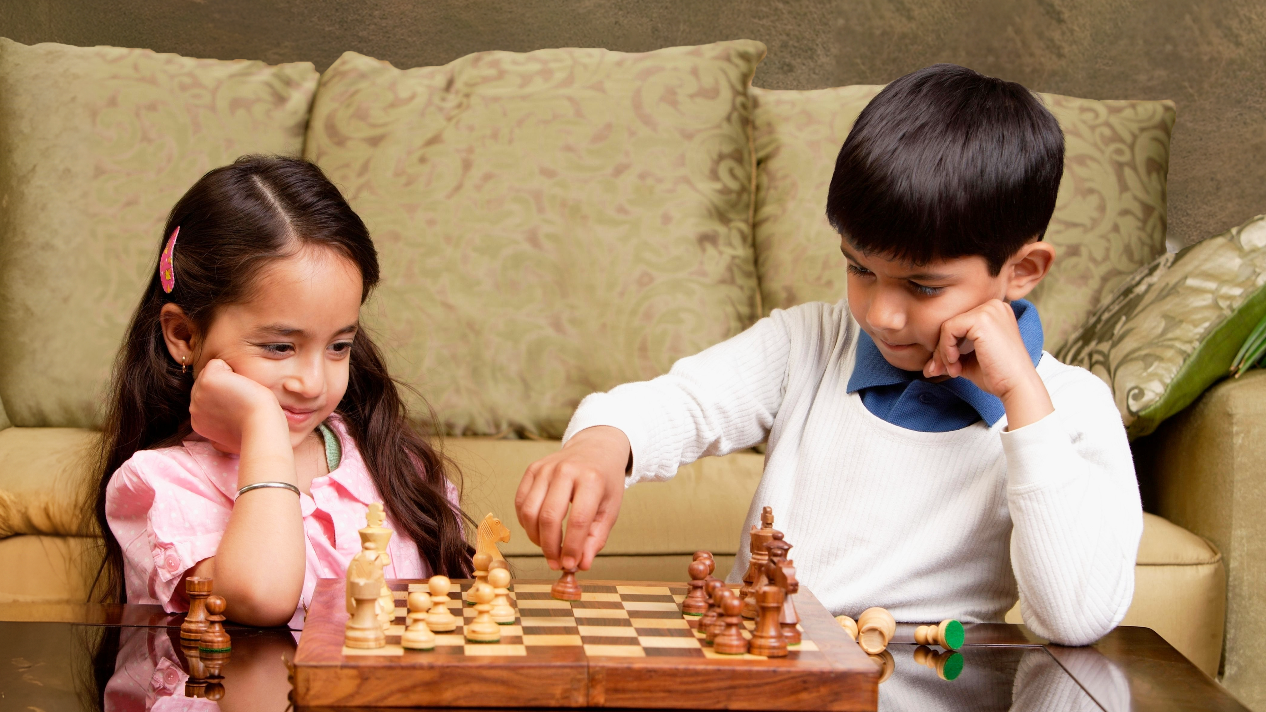 Two children play chess on a wooden board. The girl, in a pink shirt, looks thoughtful, while the boy, in a white sweater, makes a move.
