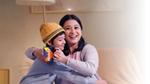 A smiling woman hugs a child in a yellow hat holding a Horlicks packet, sitting on a cozy chair.